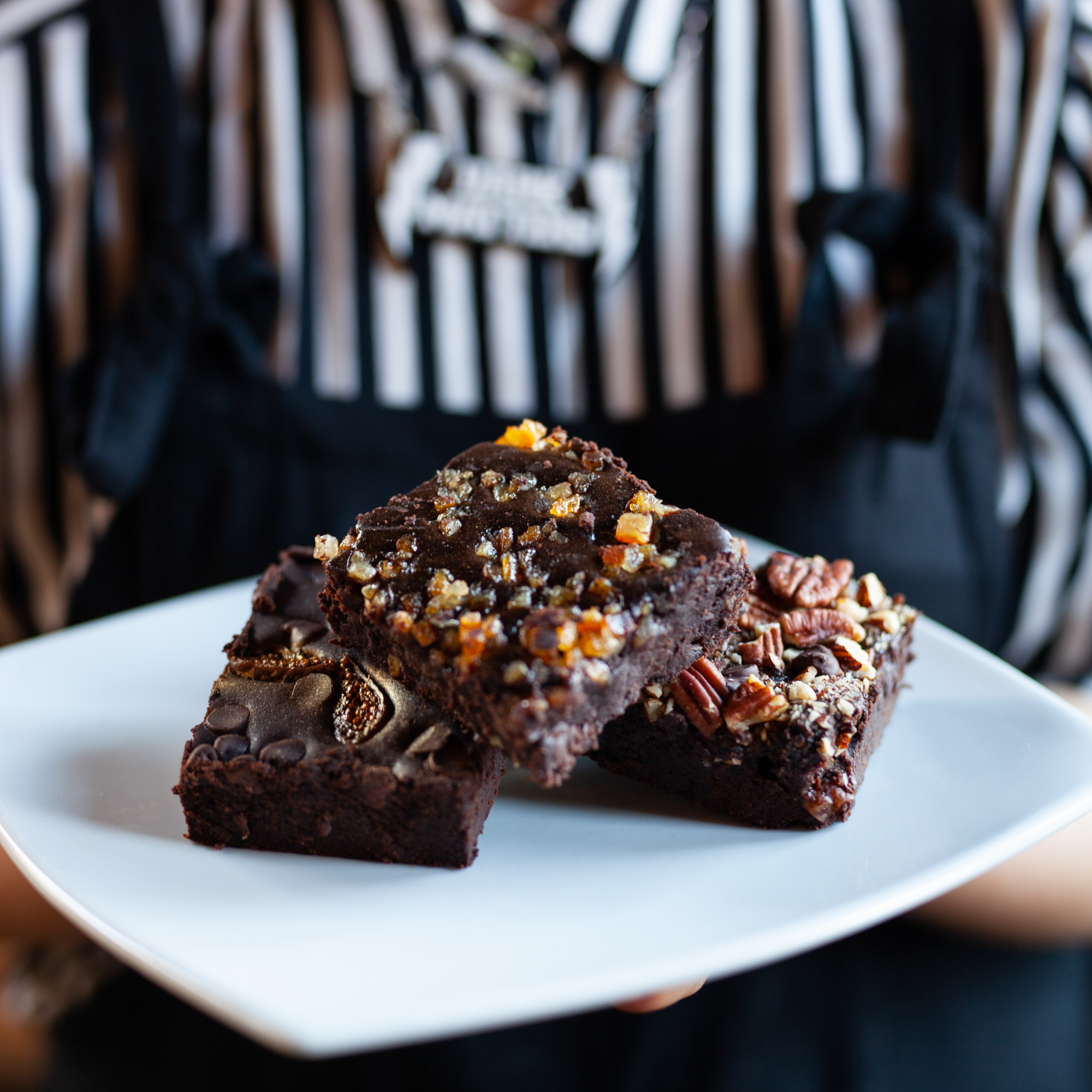 Plate of Three Vegan Brownies Held by Woman in Striped Shirt and Apron